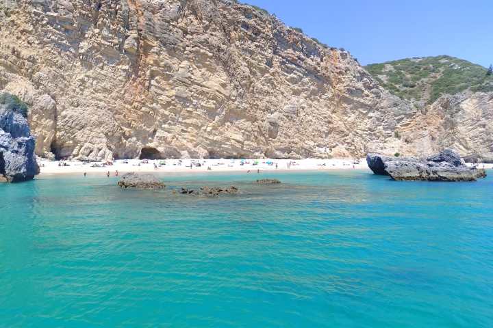 Coastal beach with clear turquoise water and rocky cliffs under a blue sky.