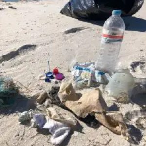 Trash and plastic debris on a sandy beach with a black garbage bag.