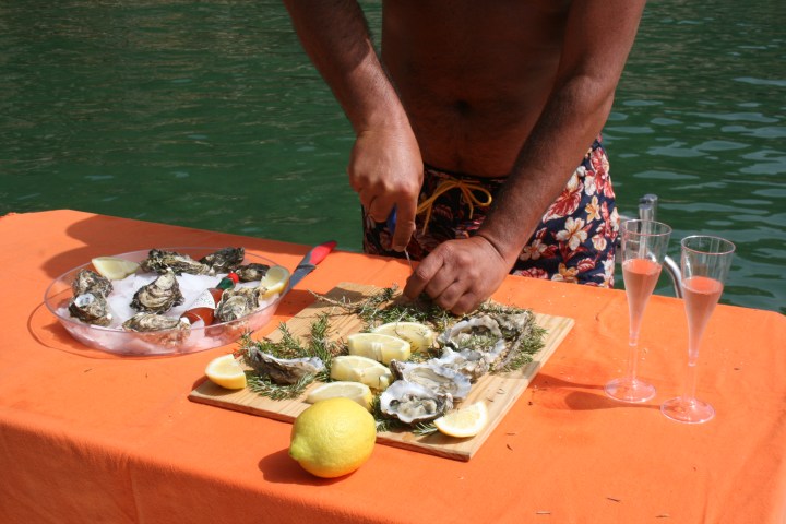 Person shucking oysters by water with citrus and two glasses on an orange tablecloth.