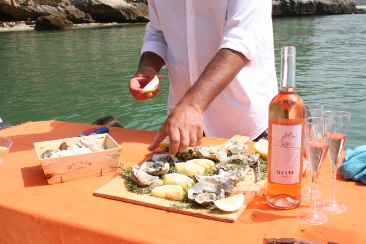 Person preparing oysters with lemon on a table by water, with a bottle of rosé wine and two glasses.