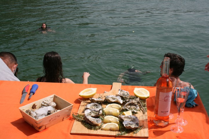 People swimming near a table with oysters, lemon slices, and a bottle of rosé wine.