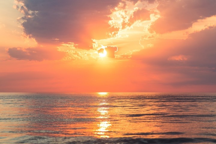 Sunset over calm ocean with clouds and orange-pink sky reflections on water.