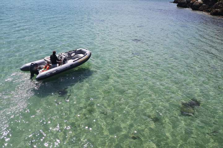 Person in a rubber boat on clear, shallow water near a rocky shore.