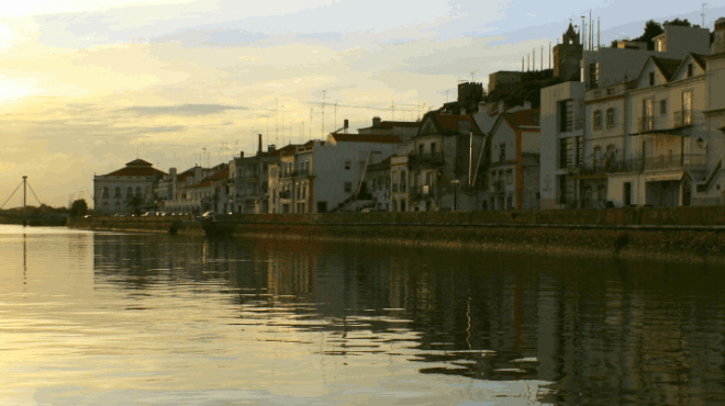 Sunset over a calm waterfront with buildings and a distant bridge.