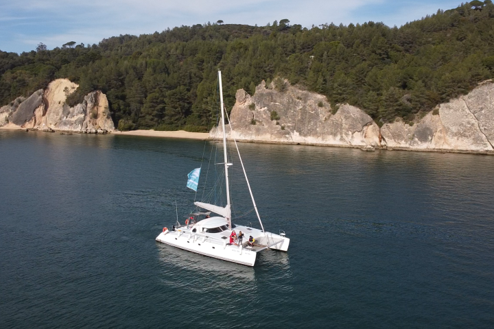 Catamaran sailing near rocky, tree-lined shore.