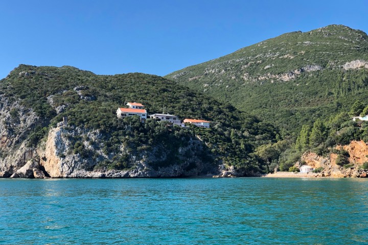 Coastal hillside with houses, surrounded by blue sea and green trees.