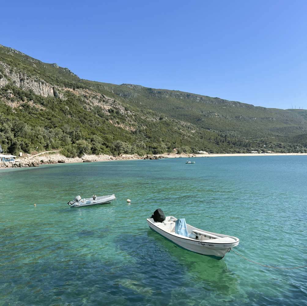 Two boats float on clear turquoise water near a green hillside under a blue sky.