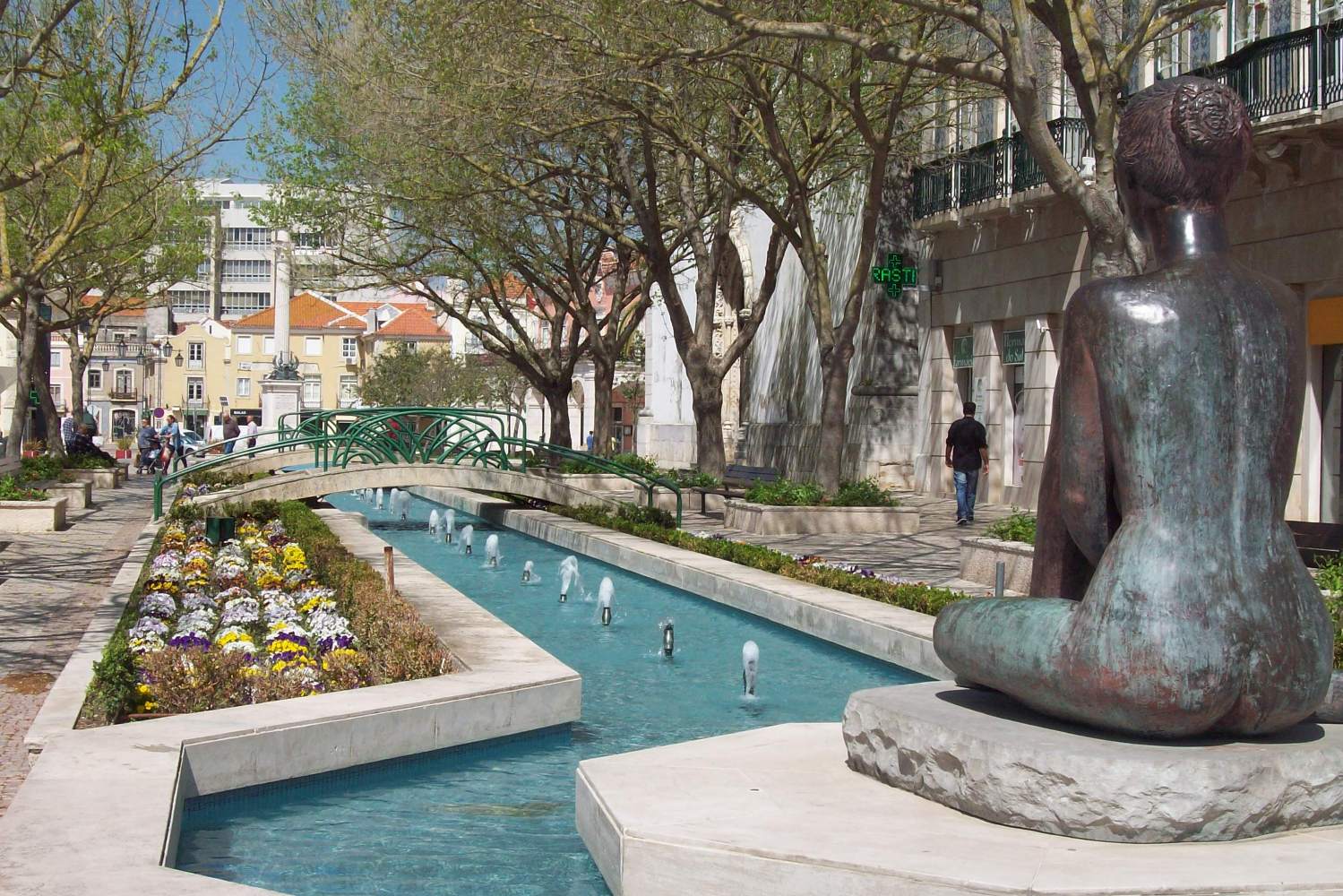 Bronze statue by a narrow canal with flowers, fountain jets, a small bridge, and people strolling nearby.