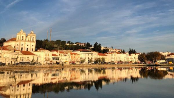 Scenic riverside town with church towers, red-roofed buildings, and reflections in water under a blue sky.