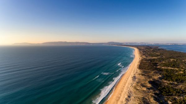Aerial view of a long sandy beach with waves and distant mountains under a clear blue sky.