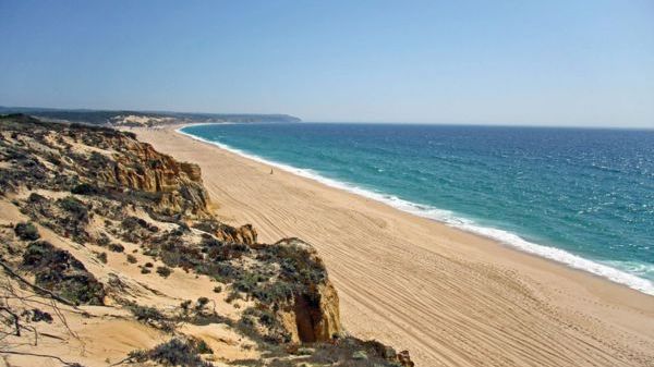 Long sandy beach with cliffs and clear blue sea under a clear sky.