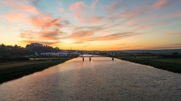 River at sunset with bridge and distant buildings under colorful sky.